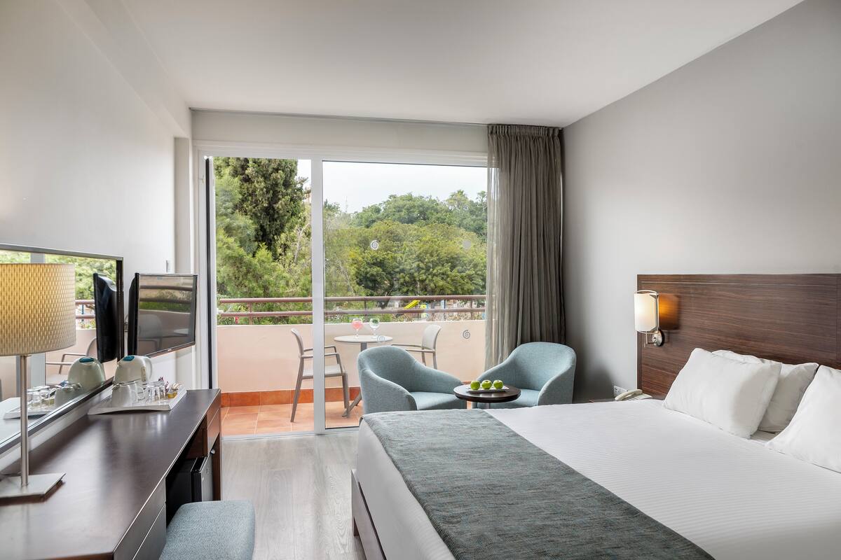 A hotel room with a bed, a desk with a TV, a chair, a small table with fruit, and a balcony with a table and chairs.