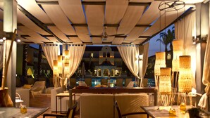 An outdoor dining area with tables and chairs, a pool in the background, and decorative lighting.