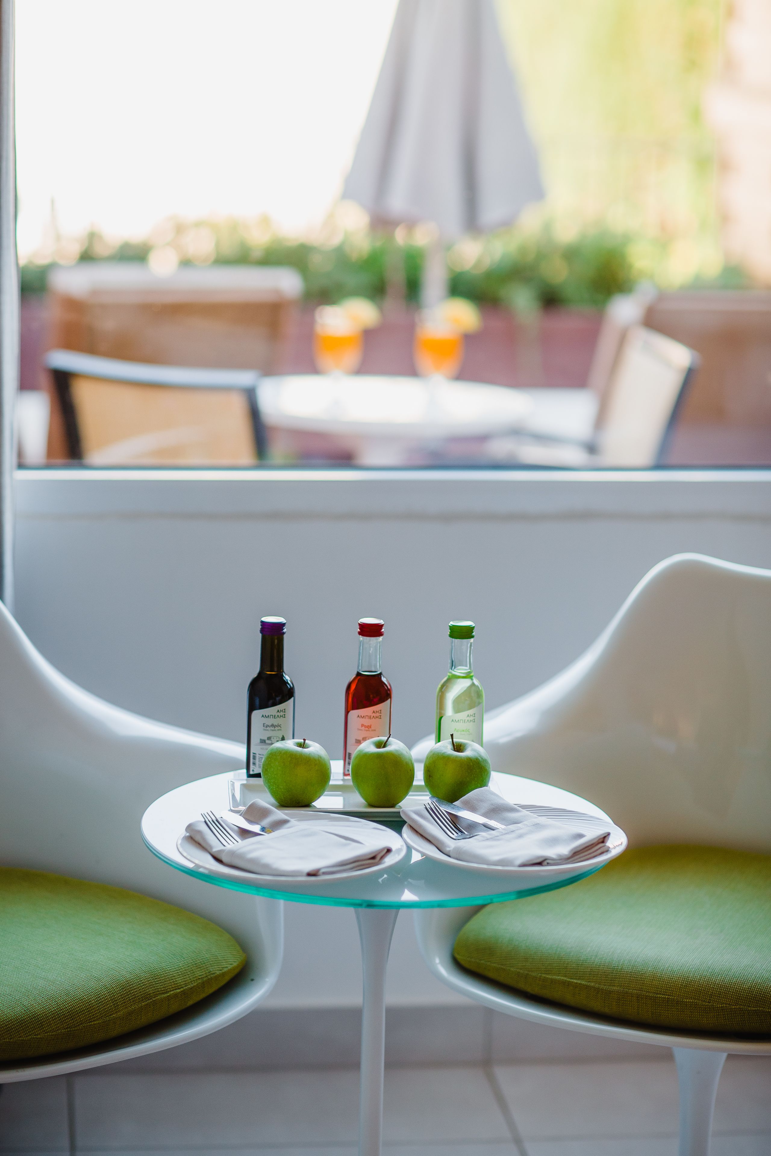 A glass table with two green chairs, a bottle of wine, two green apples, and a bottle of water.