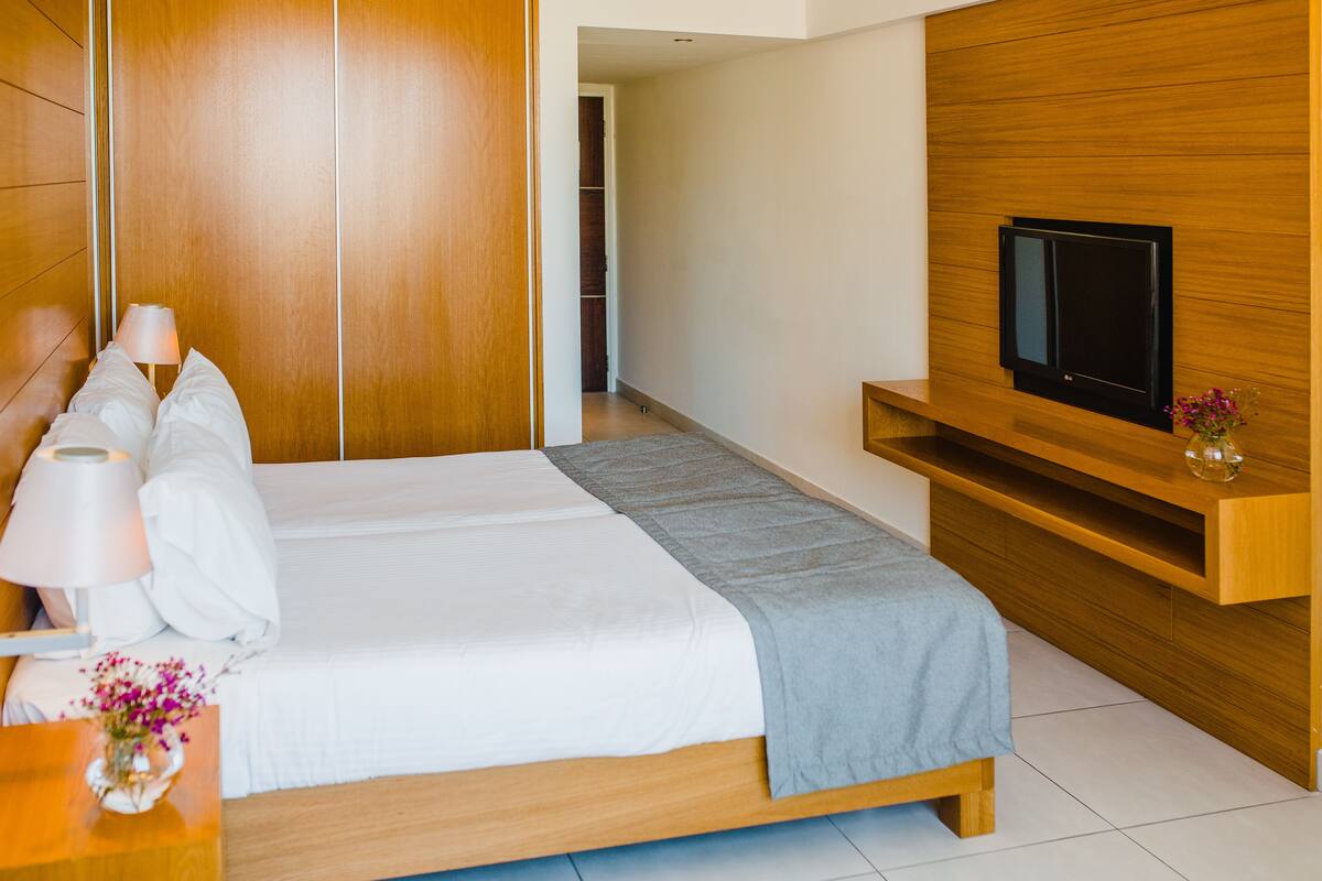 A modern hotel room with a large bed, a TV mounted on a wooden panel, and a bedside table with a lamp and flowers.