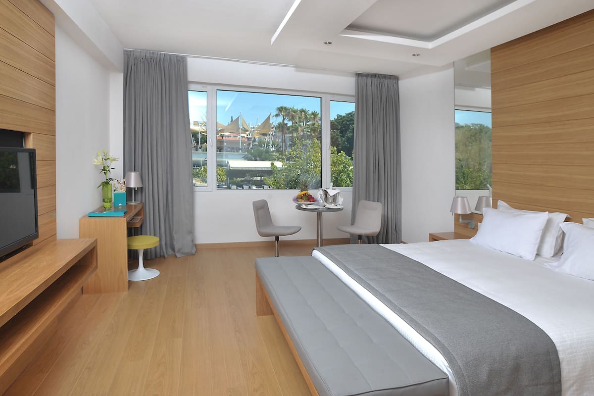 A modern hotel room with a large bed, a TV, a desk with a chair, and a view of the city.