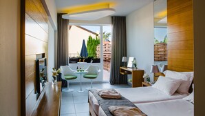 A hotel room with a bed, a desk, a chair, a small table, and a balcony with plants and a view.