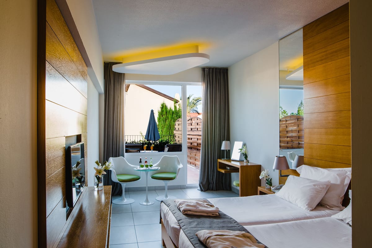 A hotel room with a bed, a desk, a chair, a small table, and a balcony with plants and a view.