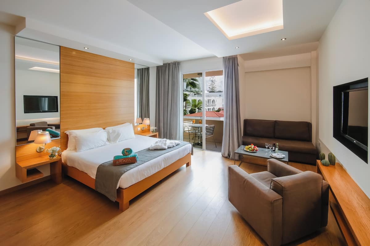 A modern hotel room with a large bed, a sofa, a TV, and a balcony with a view of the city.