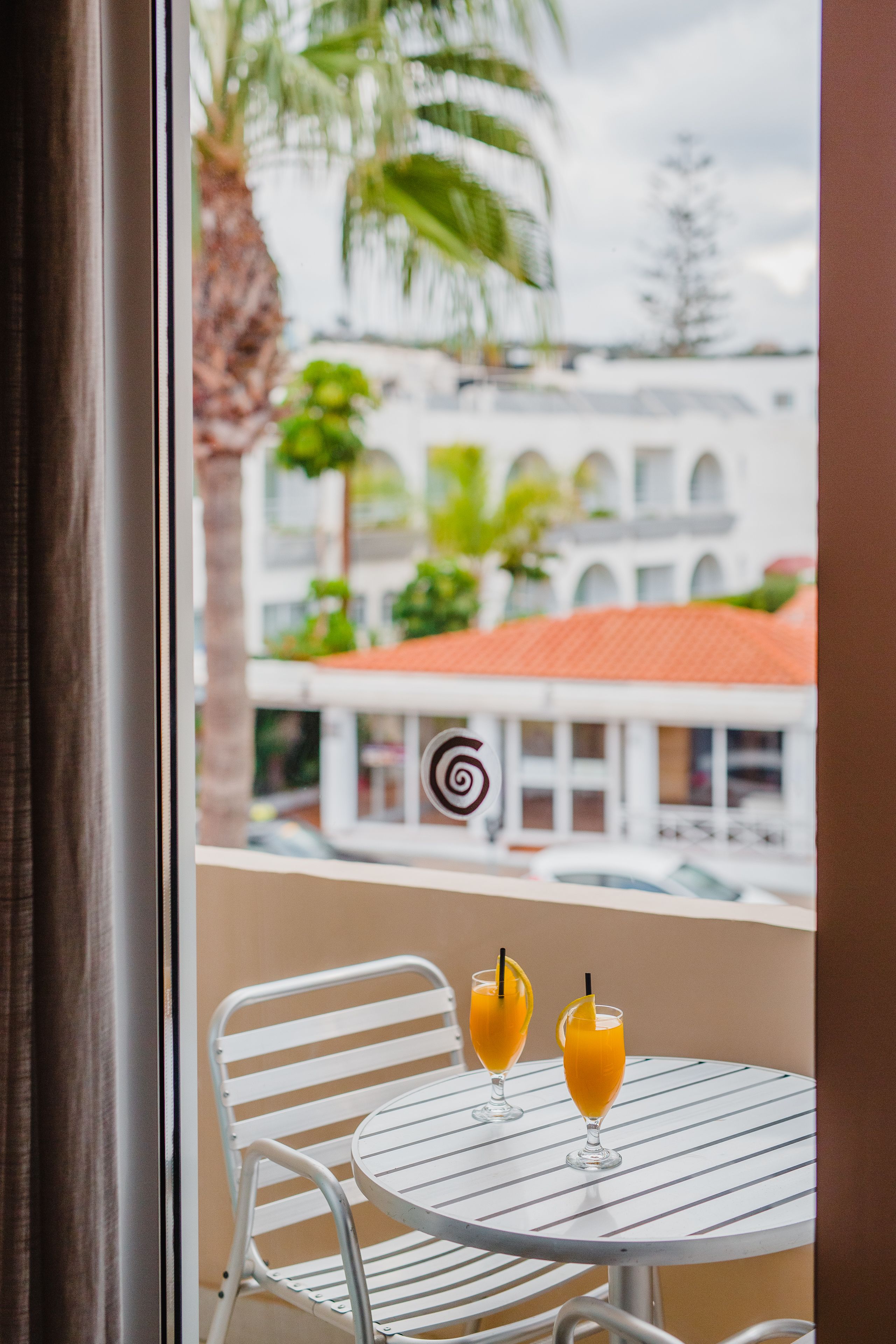 A balcony with two white metal chairs and two glasses of orange juice.