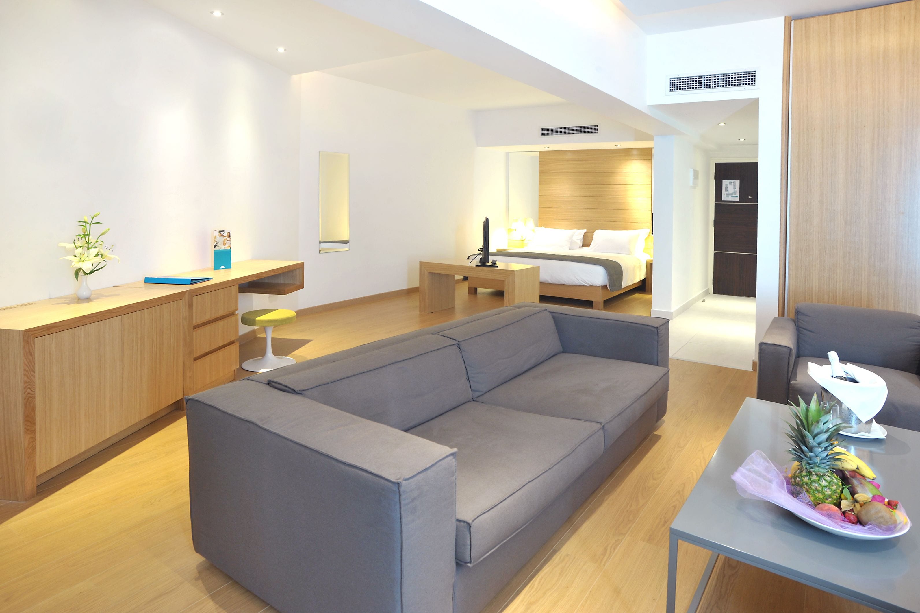 A modern hotel room with a grey sofa, a wooden desk, a bed with a wooden headboard, and a small table with fruits.