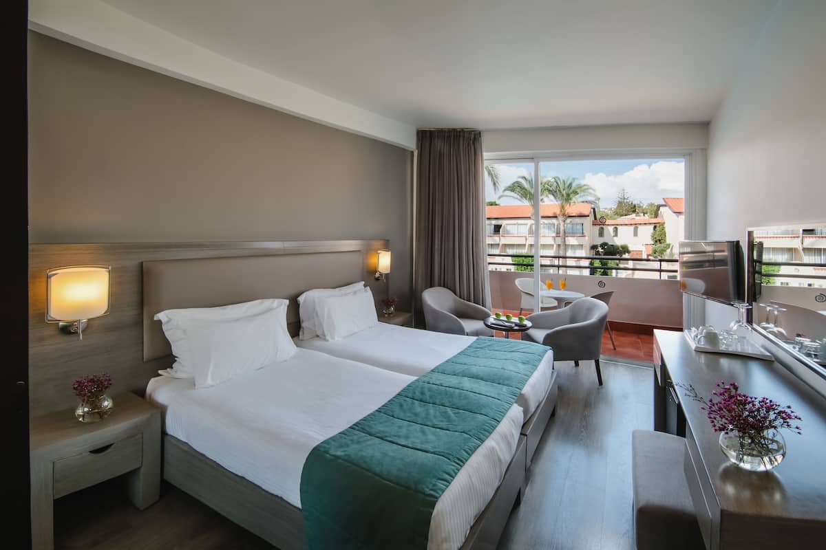 A hotel room with a bed, bedside tables, a desk, a chair, and a balcony with a view of buildings and palm trees.