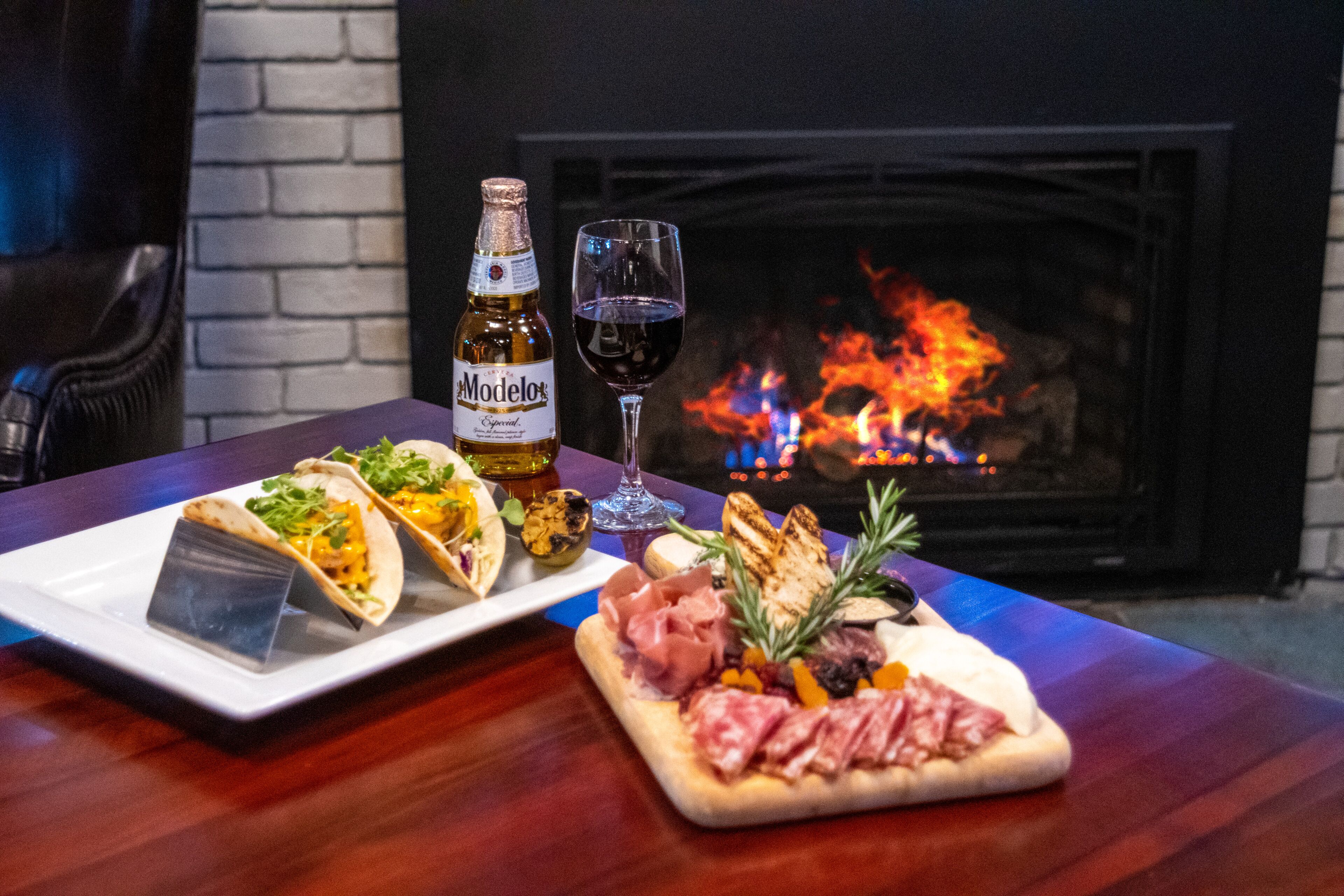 A table with a bottle of Modelo beer, a glass of red wine, and a variety of food including tacos, grilled meat, and a small pizza.