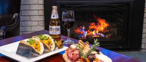 A table with a bottle of Modelo beer, a glass of red wine, and a variety of food including tacos, grilled meat, and a small pizza.