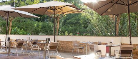 An outdoor patio with tables and chairs, shaded by large umbrellas, set against a backdrop of lush greenery.