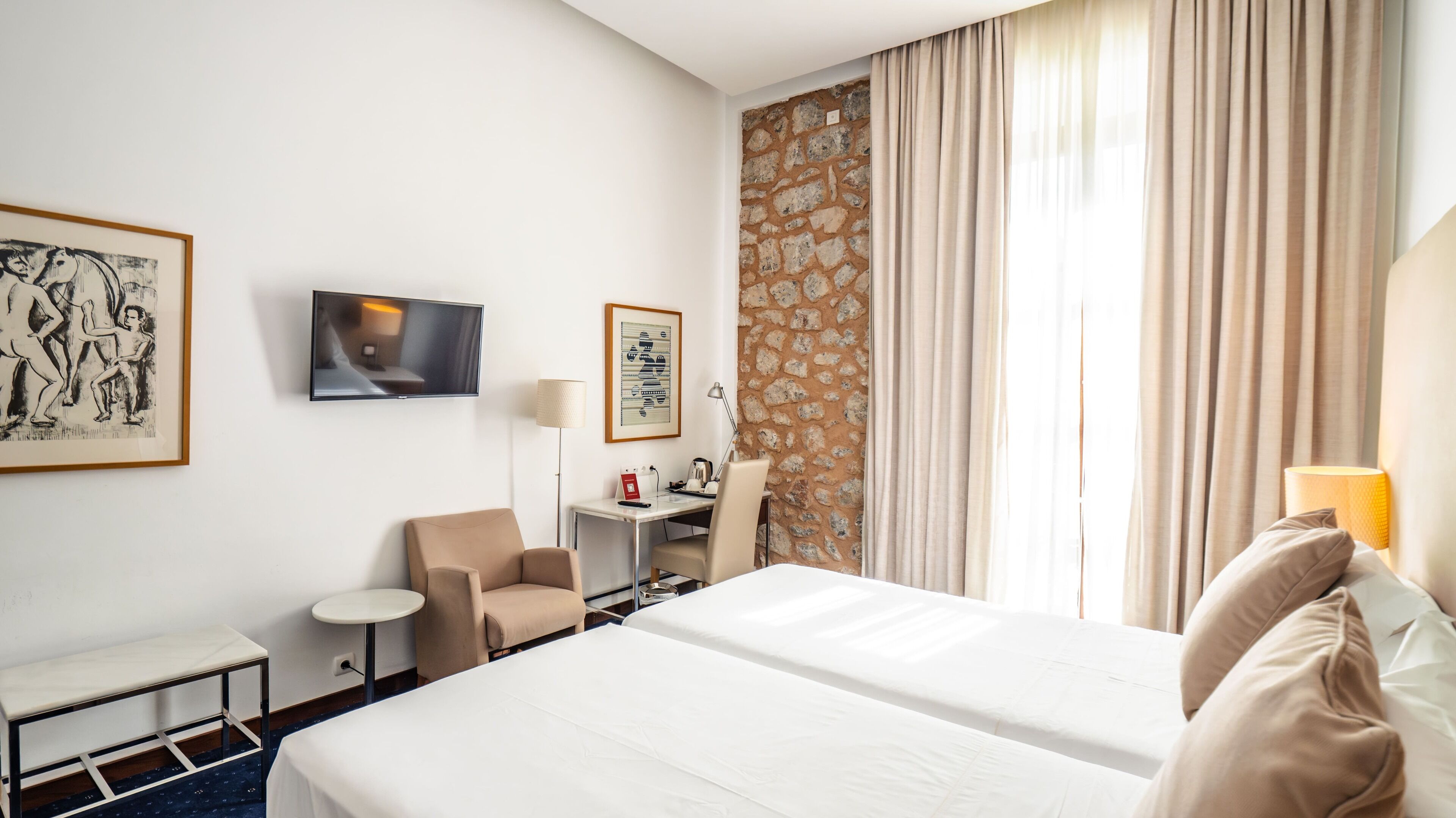 Una habitación de hotel con cama, escritorio, silla, televisor y obras de arte en las paredes.