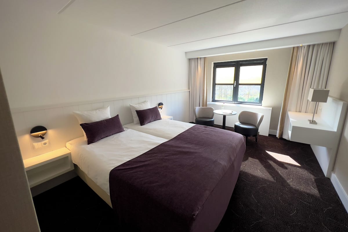Een hotelkamer met een groot bed, twee nachtkastjes, een kleine ronde tafel met stoelen, een raam met gordijnen en een bijzettafeltje met een lamp.