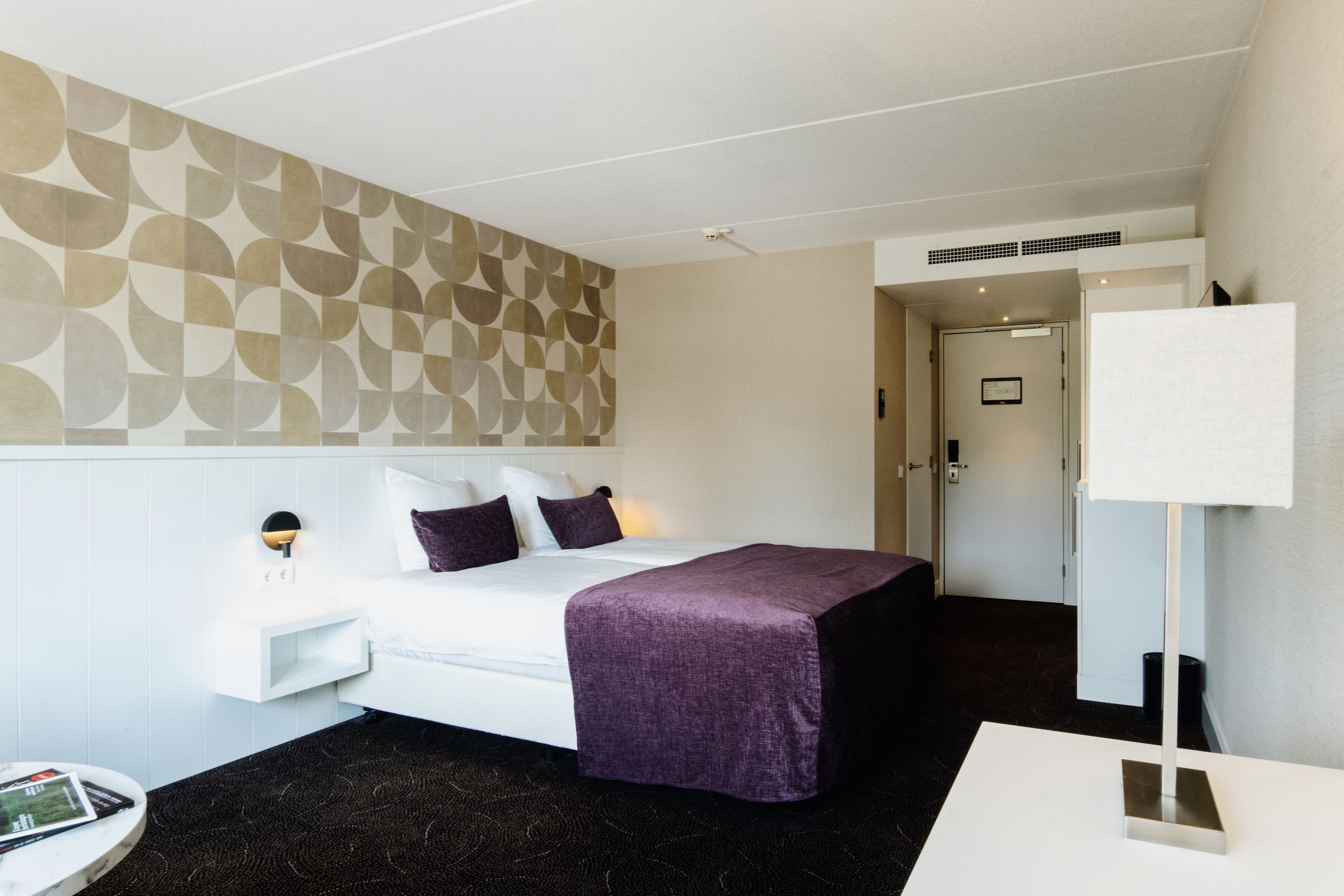 Une chambre d’hôtel avec un lit, une table de chevet, une lampe et un mur orné d’un motif géométrique.