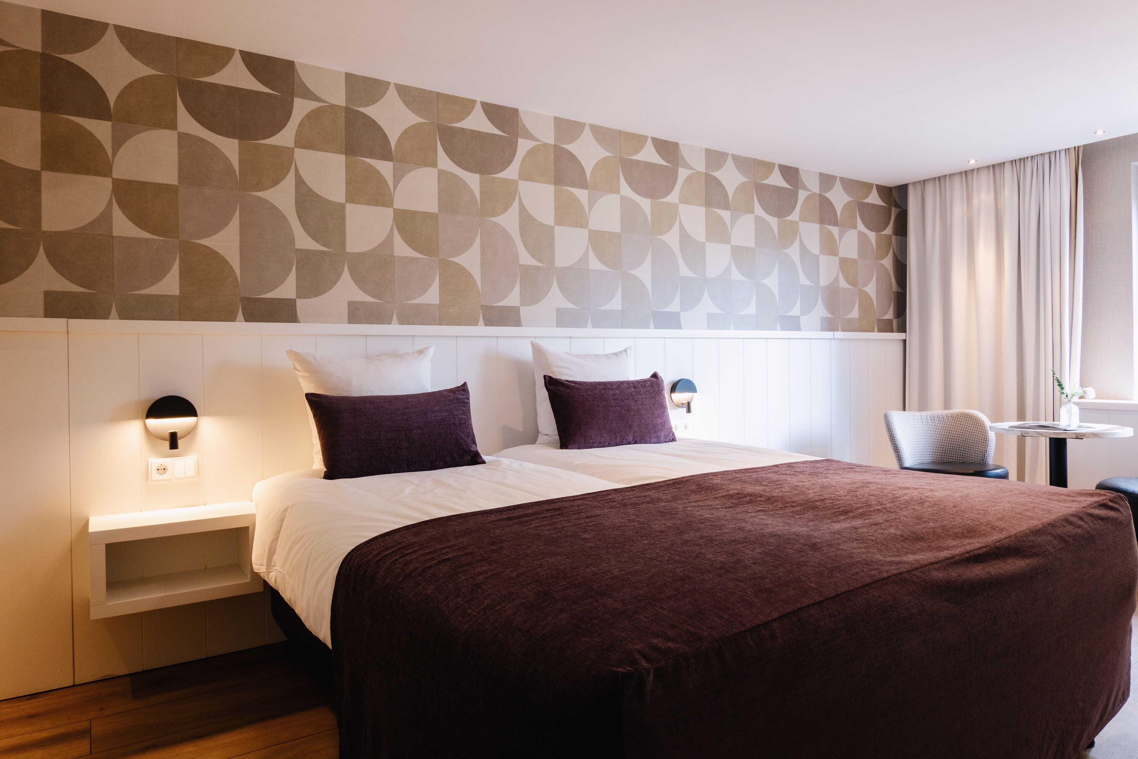 A hotel room with a large bed, a bedside table with a lamp, a chair, a small table, and a window with curtains.