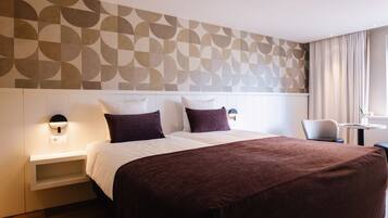 A hotel room with a large bed, a bedside table with a lamp, a chair, a small table, and a window with curtains.