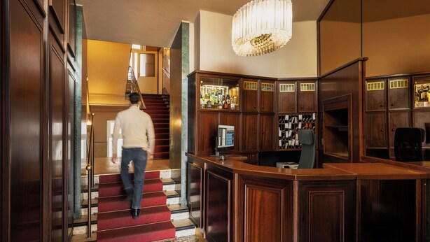 Um saguão de hotel com uma escadaria imponente, uma mesa de recepção e um espaço de bar.