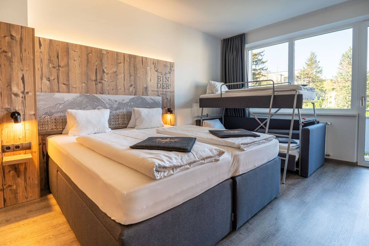 Una habitación de hotel moderna con una cama grande, una cama litera y vistas a árboles a través de un ventanal.