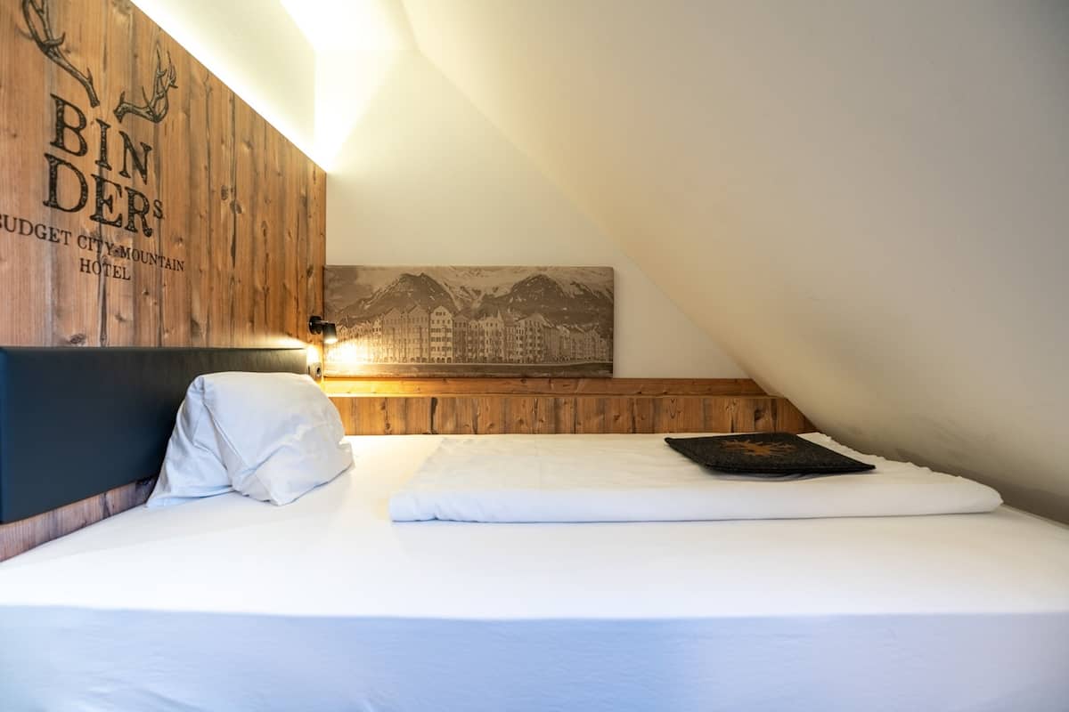 Una cama bien hecha con ropa blanca, un cabecero de madera con el texto "BIN DERS BUDGET CITY MOUNTAIN HOTEL" y una fotografía enmarcada de montañas en la pared.