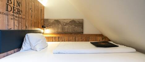A neatly made bed with white linens, a wooden headboard with the text "BIN DERS BUDGET CITY MOUNTAIN HOTEL", and a framed picture of mountains on the wall.