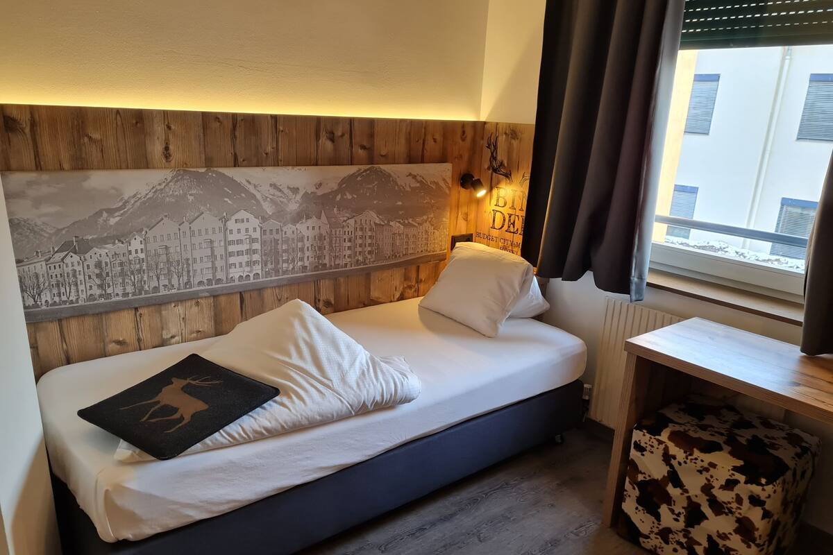 Un dormitorio con cama, cabecera de madera, una obra de arte urbana, una mesita pequeña y una ventana con cortinas.