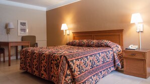 A hotel room with a large bed, two bedside lamps, a wooden nightstand, a desk with a chair, and a framed picture on the wall. - America's Inn - Houston (Houston)