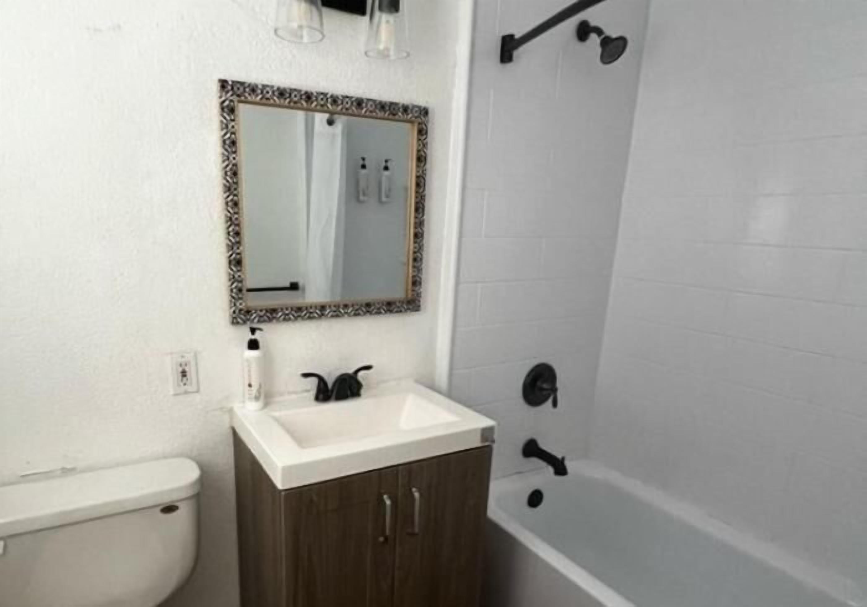 Ein Badezimmer mit Toilette, einem Holzkörperschen mit weißem Waschbecken, einem Spiegel mit dekorativem Rahmen und einer Badewanne mit schwarzem Wasserhahn.