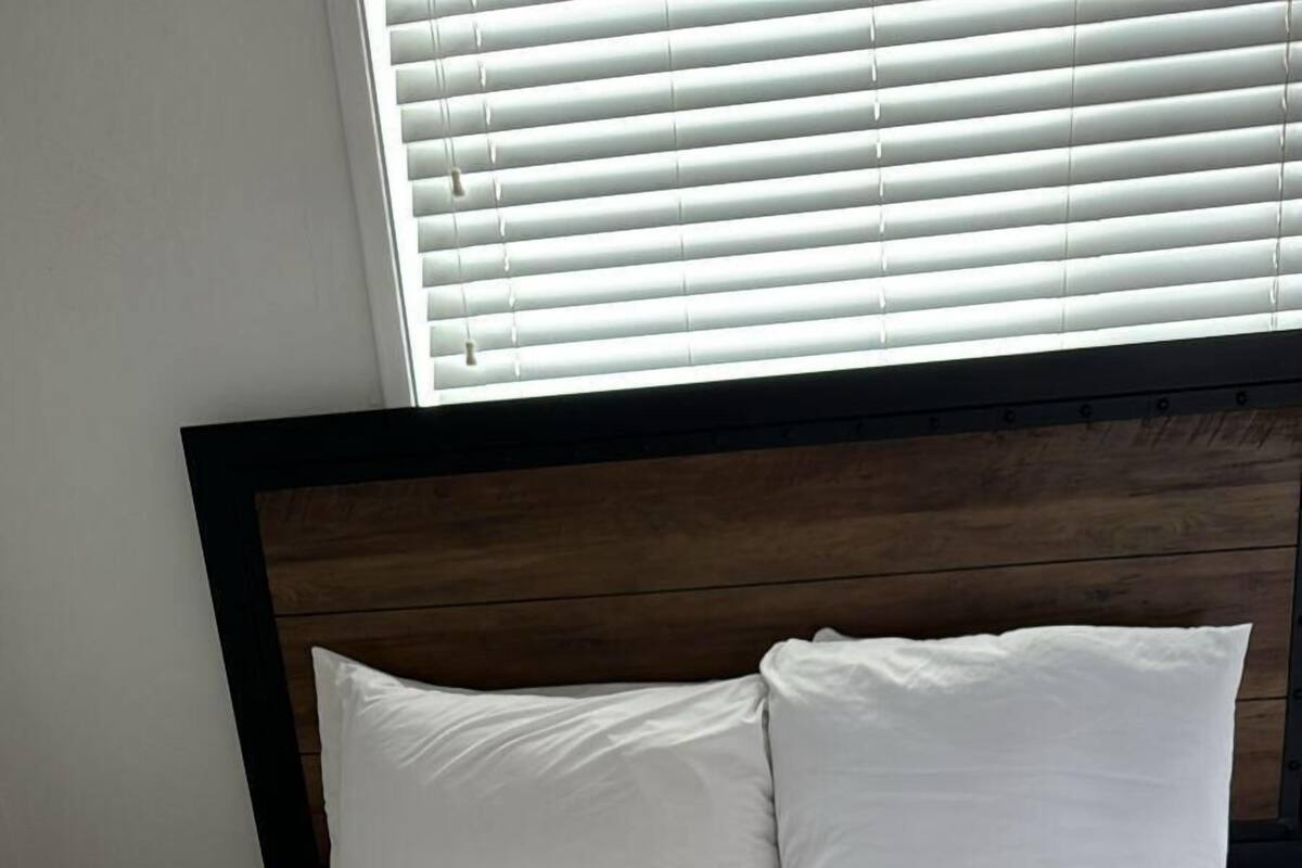A neatly made bed with white linens and rolled towels, a wooden headboard, and white blinds on the window.