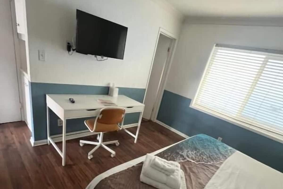 A bedroom with a bed, a desk, a chair, a television, and a window with blinds.