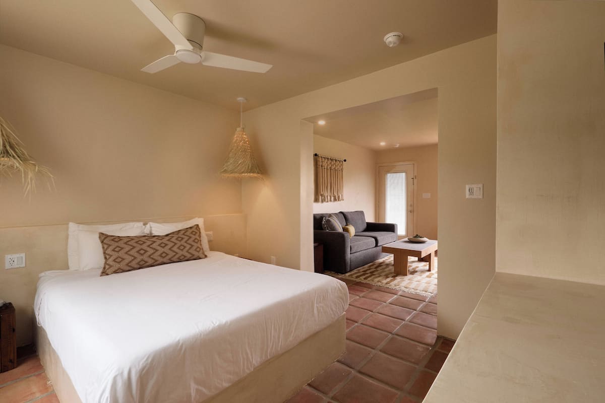 A bedroom with a bed, a ceiling fan, a hanging light fixture, and a view into a living area with a sofa and a coffee table.
