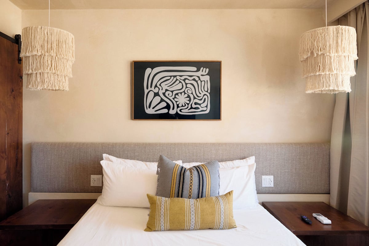 A hotel room with a bed, two bedside tables, a wall-mounted artwork, and two hanging light fixtures.