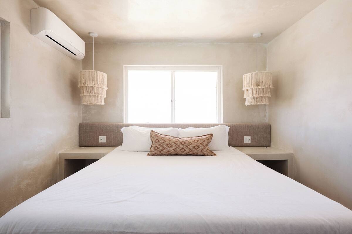 A bedroom with a large bed, two bedside lamps, a window, and an air conditioning unit.
