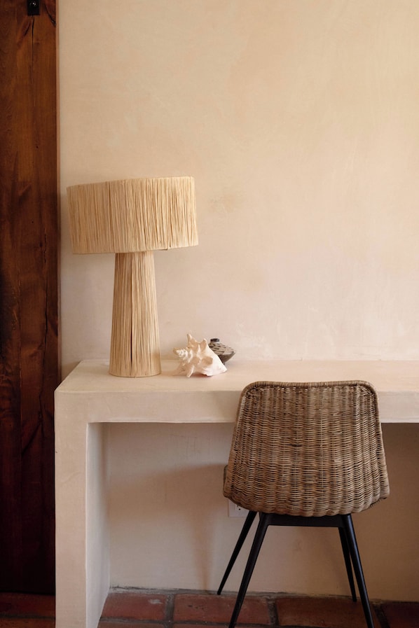 A room with a wooden door, a lamp on a white shelf, a wicker chair, and a small decorative item.