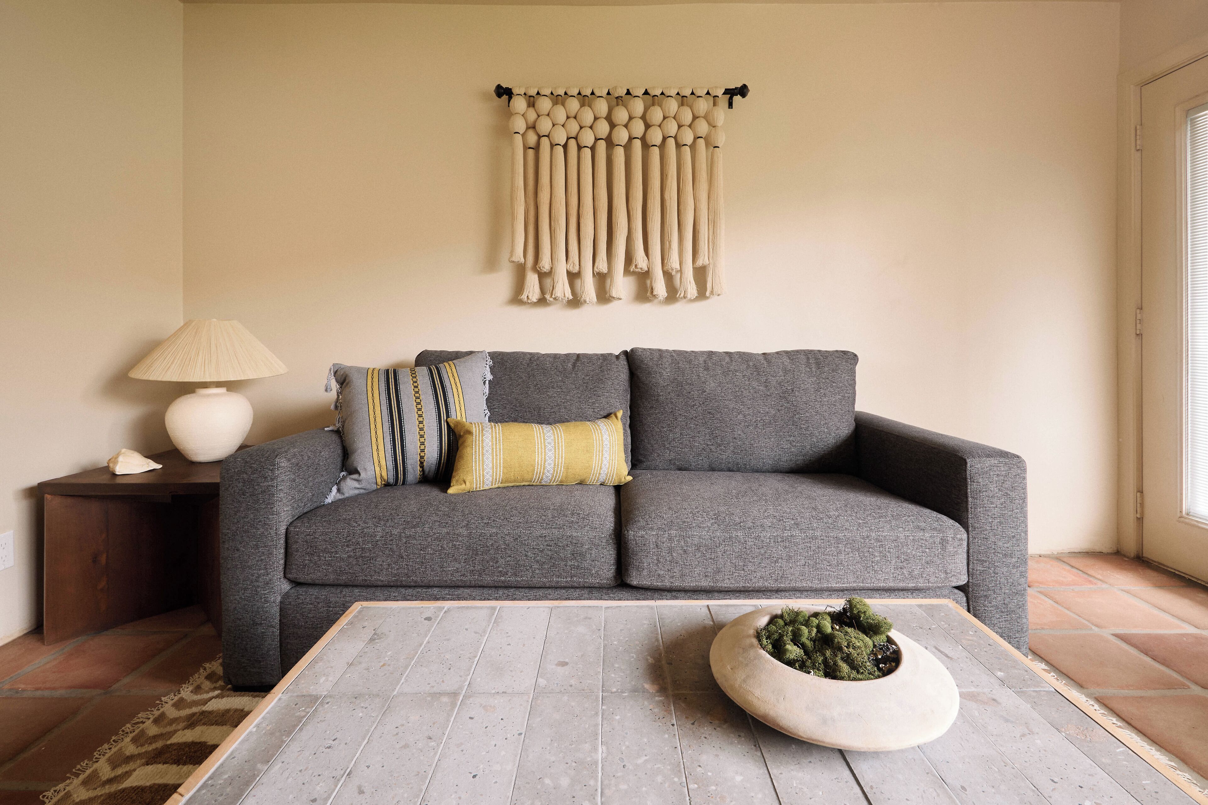 A living room with a grey sofa, a wooden coffee table with a plant, a lamp, and a wall hanging.