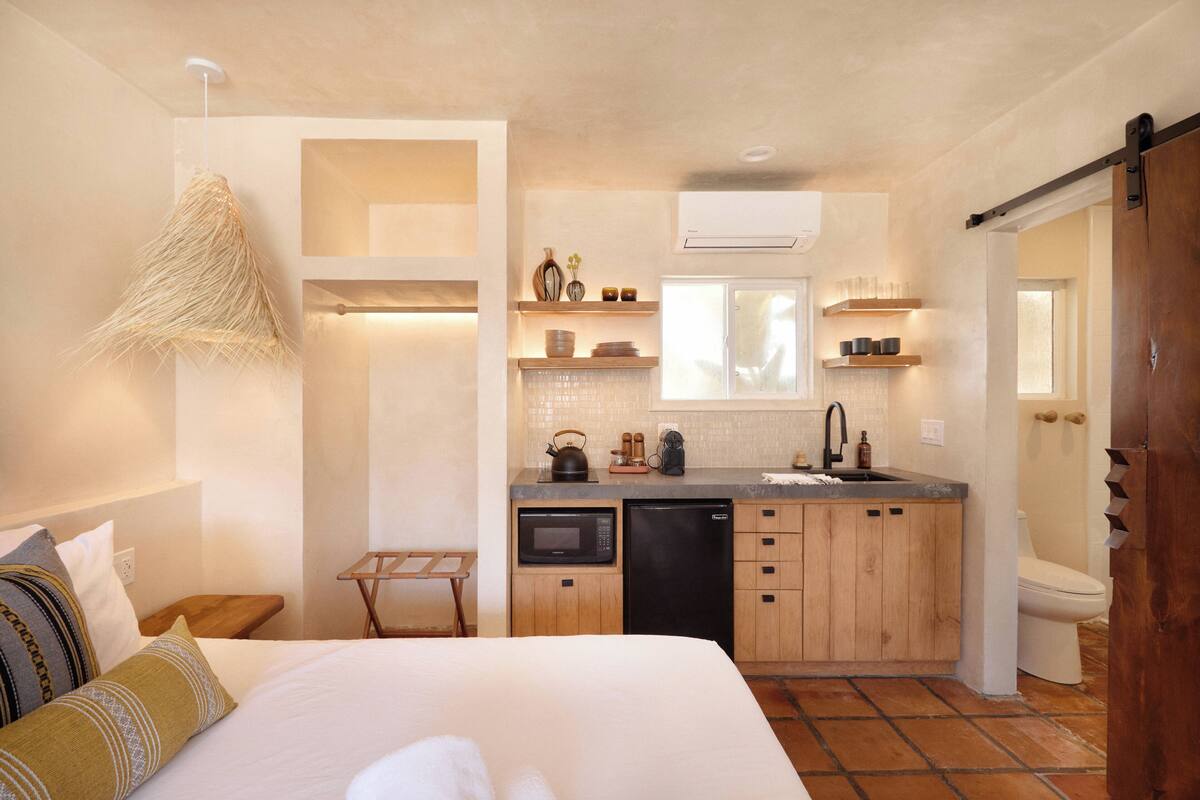 A compact hotel room with a bed, a small table, a kitchenette with a microwave, a sink, and a toilet.