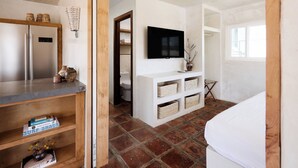A bedroom with a bed, a TV mounted on the wall, a wooden shelf with storage baskets, and a tiled floor. - Los Bungalows (Imperial Beach)