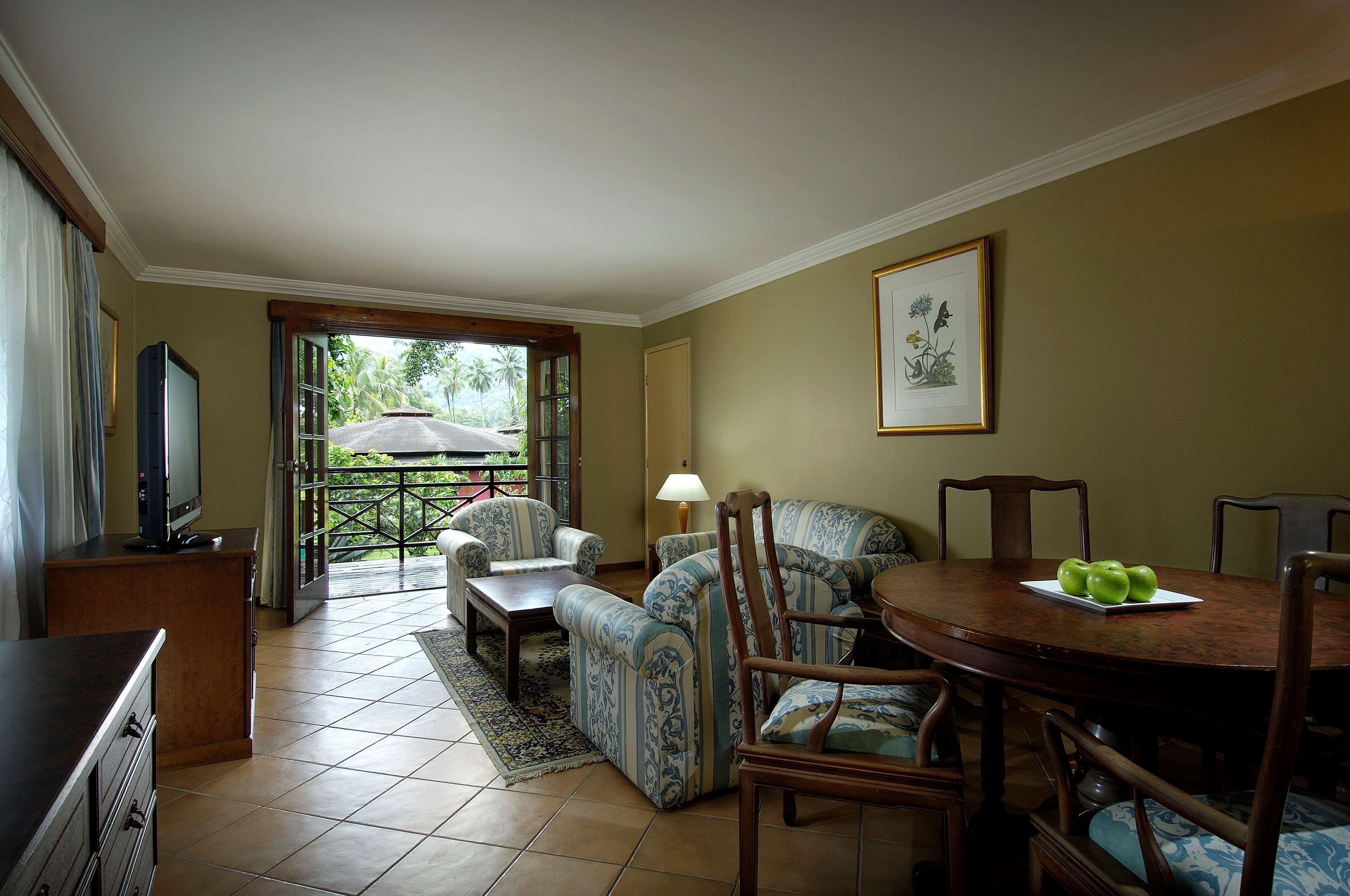 Sala de estar com televisão, sofá, cadeiras, uma mesa de jantar com maçãs e vista para uma estrada e vegetação através de uma porta de vidro.