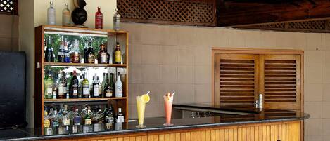 A pool bar with wooden bar counter, shelves of bottles, two wicker bar stools, and a ceiling fan.