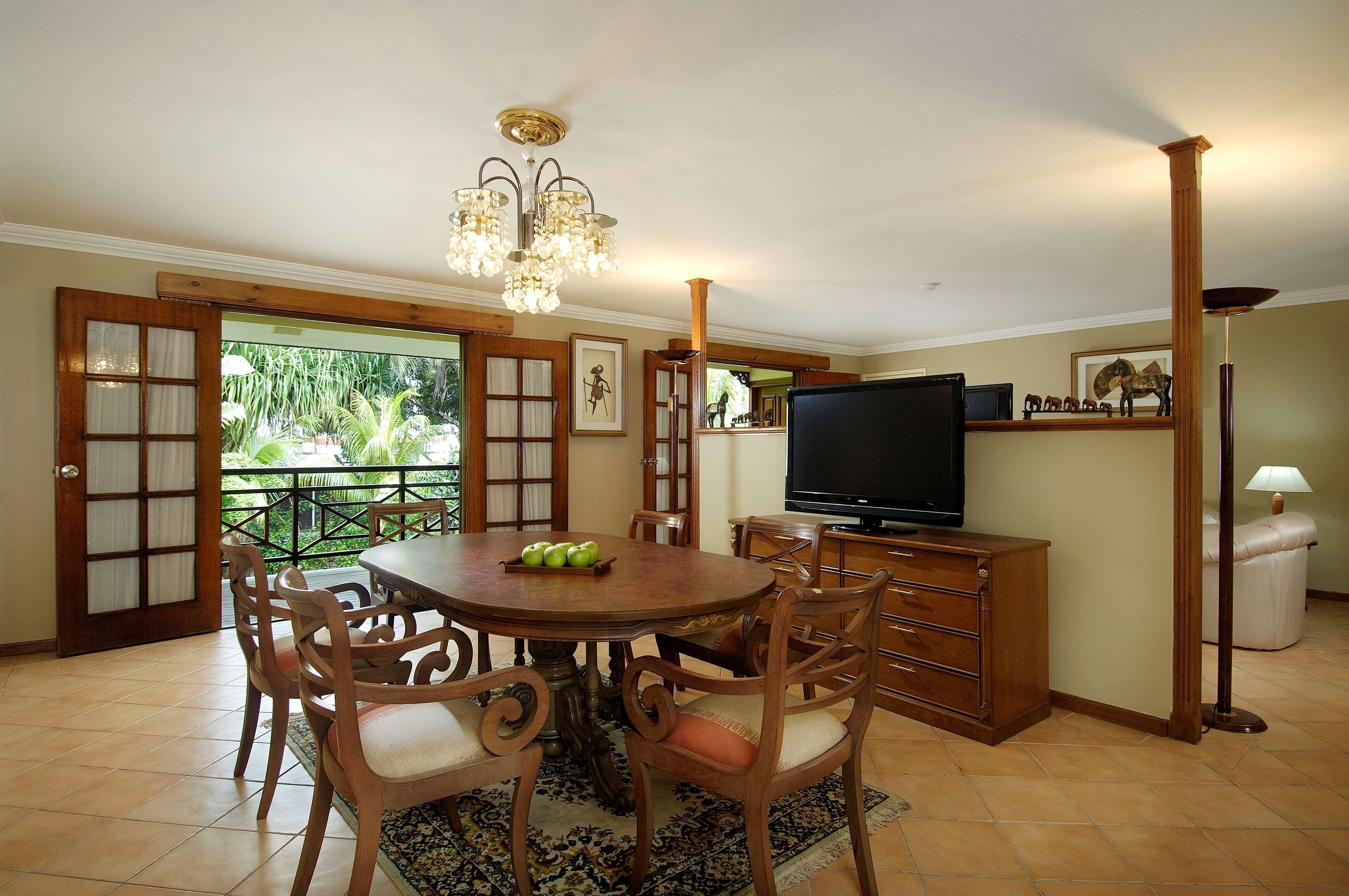 A dining area with a wooden table, chairs, and a chandelier. There is a television on a stand and a window with a view of greenery.