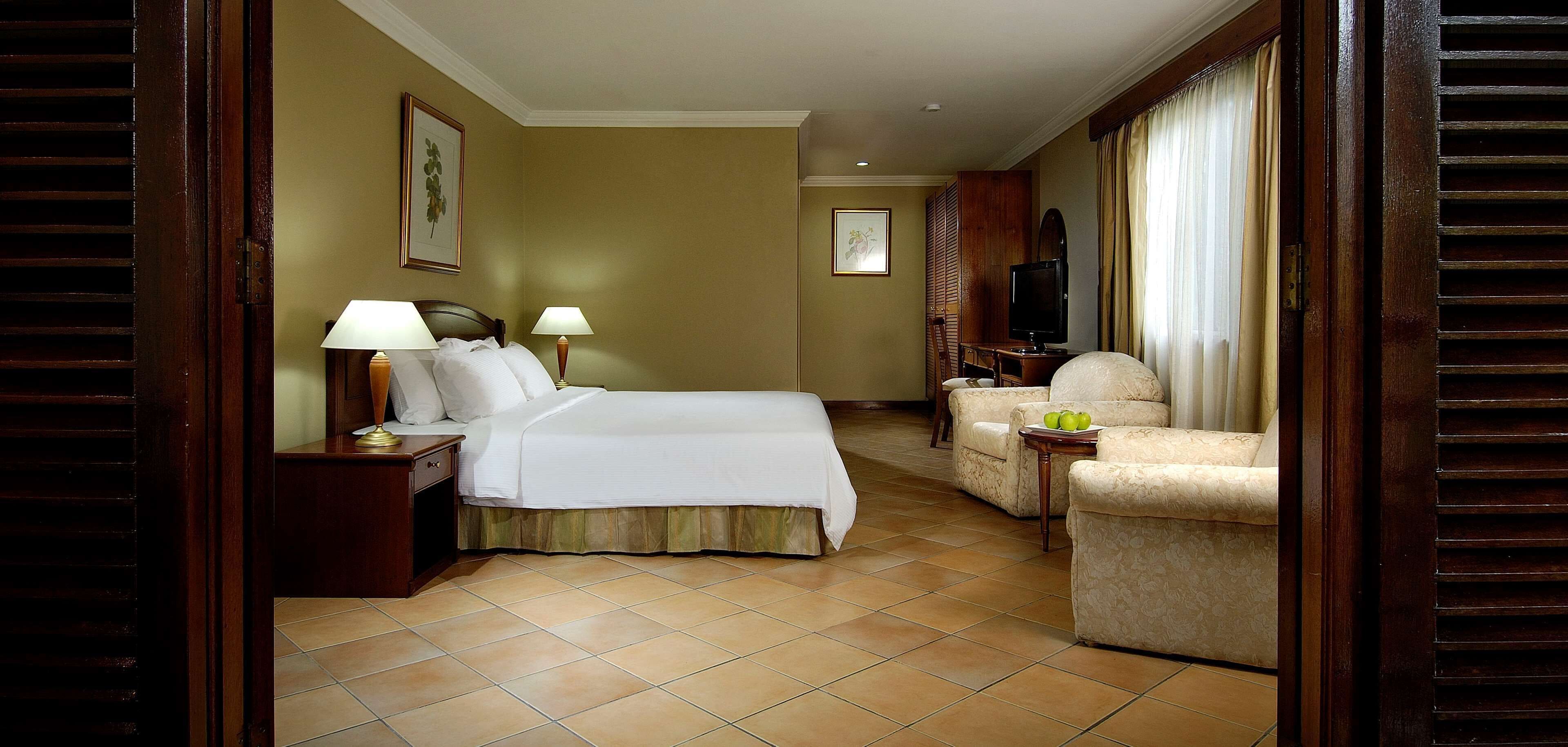 Une chambre d’hôtel avec un lit, deux tables de chevet avec des lampes, une télévision, un canapé et une petite table avec des fruits.