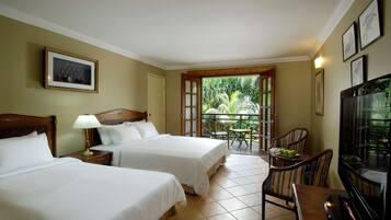 A hotel room with two beds, a TV, a dining table with chairs, and a balcony with a view of greenery.