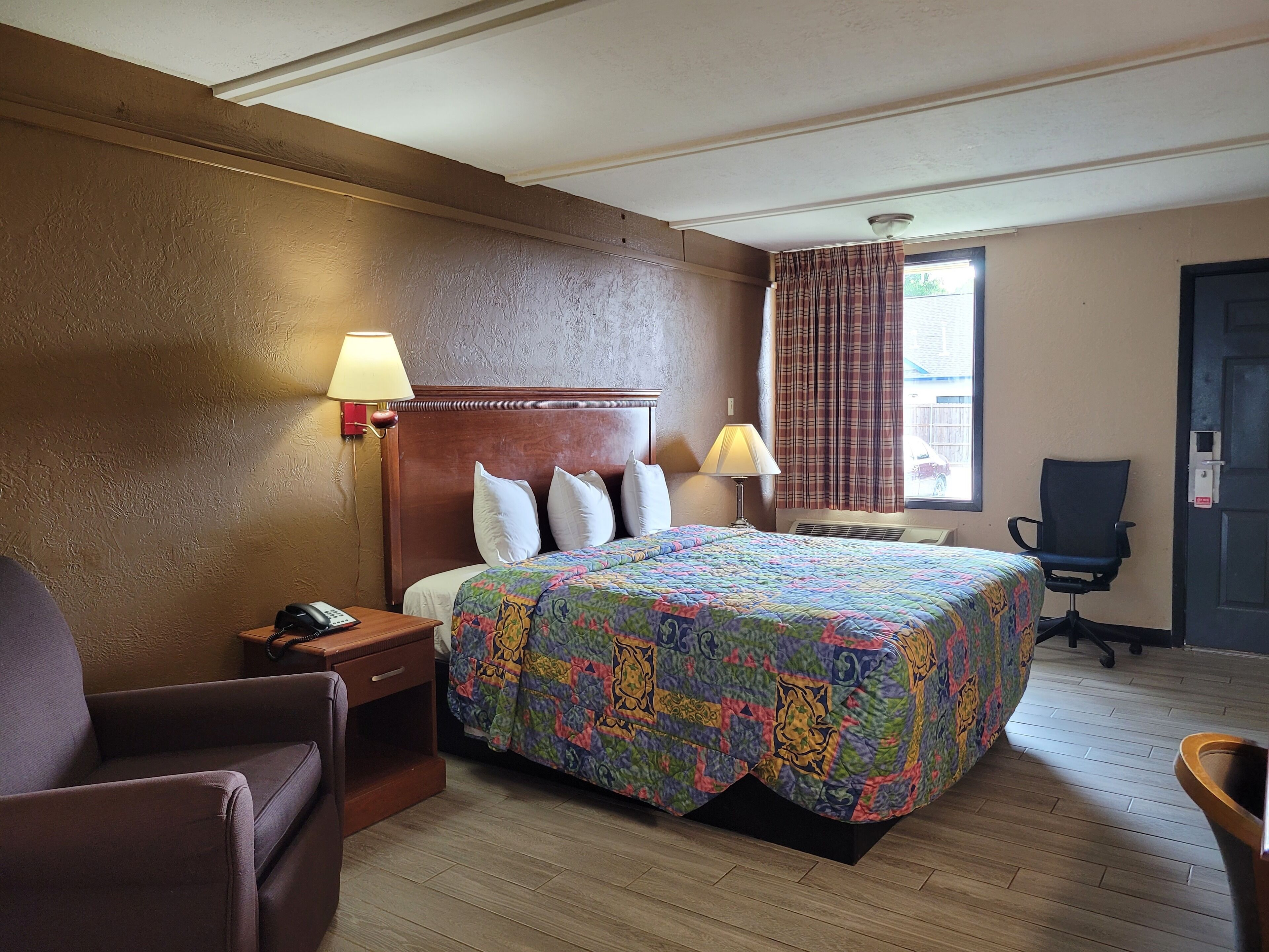 A hotel room with a wooden headboard, a bed with a colorful quilt, two bedside lamps, a chair, a desk with a phone, and a window with curtains.