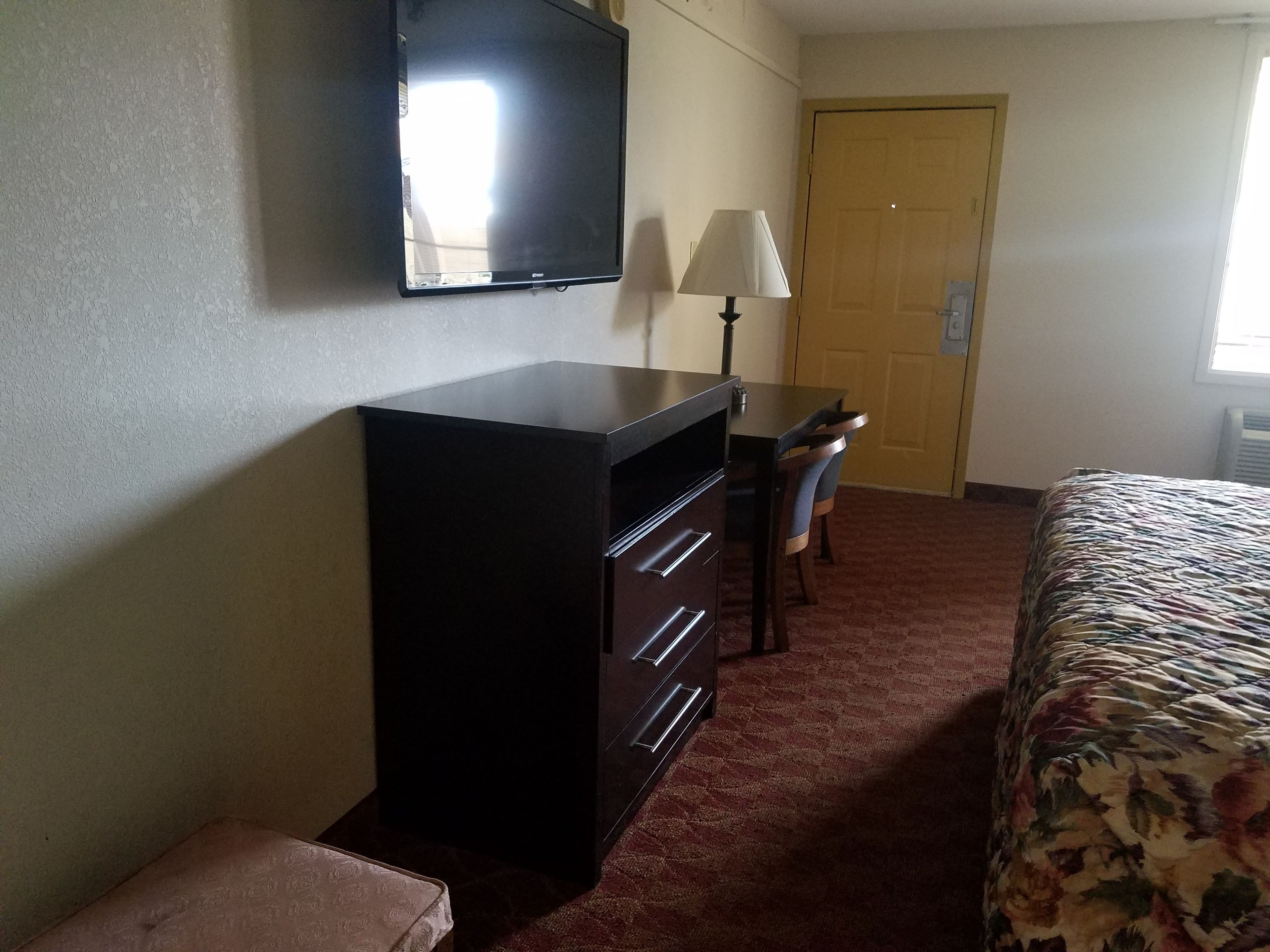 A hotel room with a bed, a desk with a lamp, a television on the wall, and a door.
