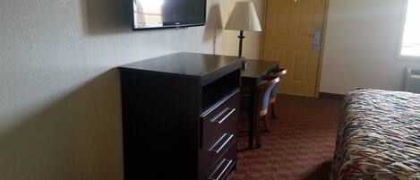 A hotel room with a bed, a desk with a lamp, a television on the wall, and a door.