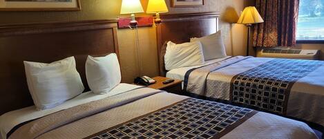 A hotel room with two beds, each with a patterned bedspread and white pillows. There are two wall-mounted lamps, a framed painting above the beds, and a window with curtains.