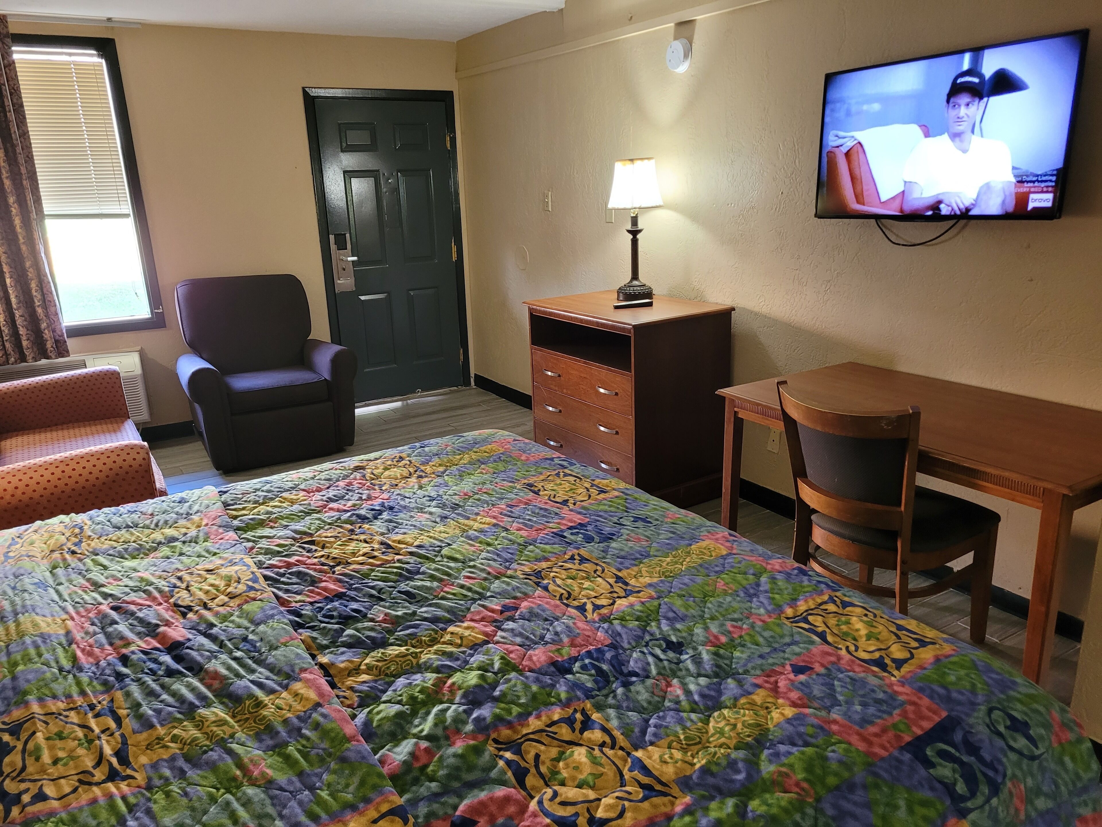 Habitación de hotel con cama, escritorio, silla, cómoda, lámpara y televisión en la pared.