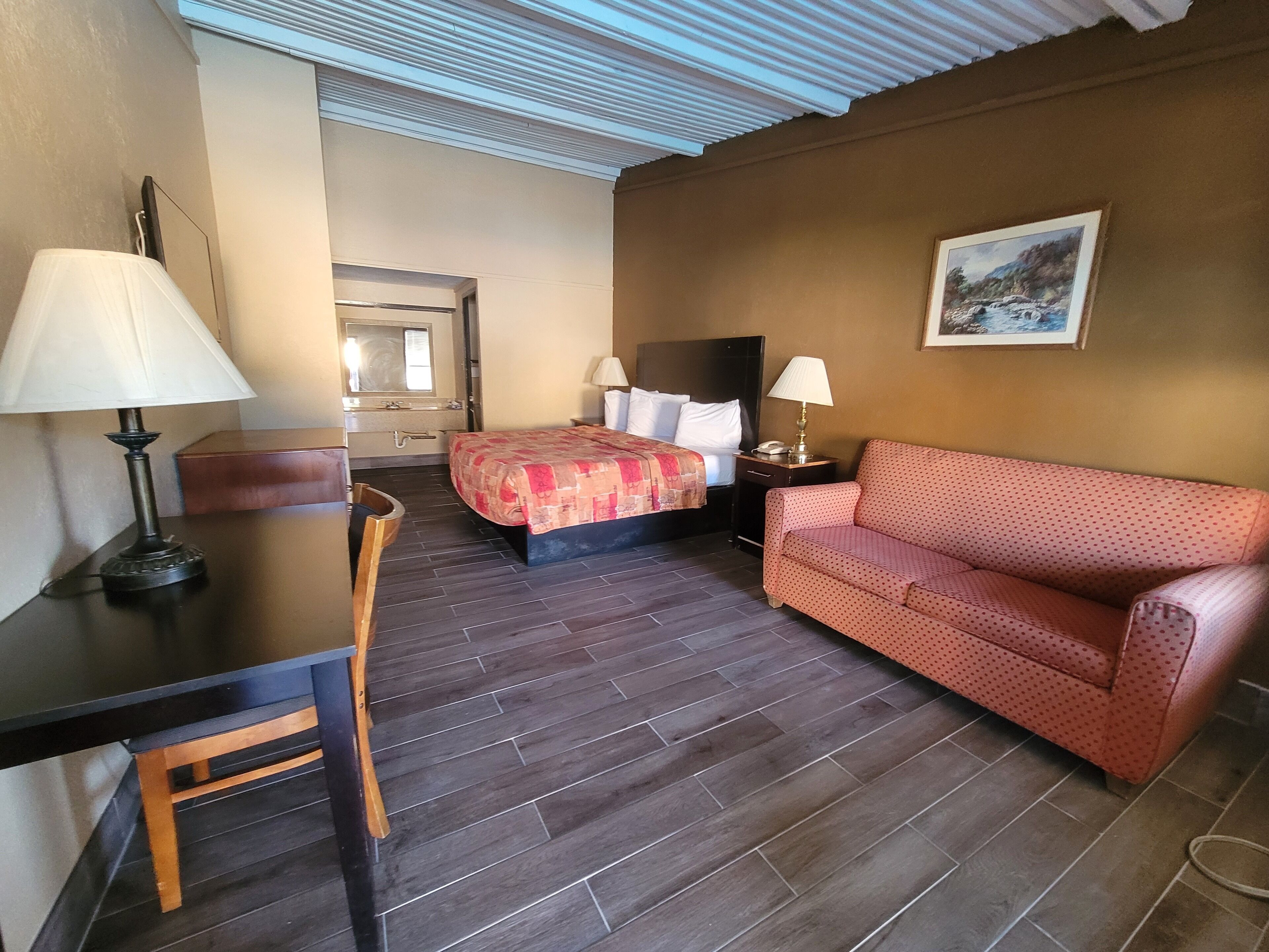 A hotel room with a bed, a desk with a lamp, a red sofa, a nightstand with a lamp, a painting on the wall, and a bathroom visible through an open door.