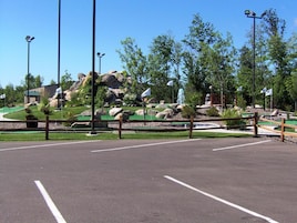 A parking lot with a green golf course area, surrounded by trees and a building with a green roof.