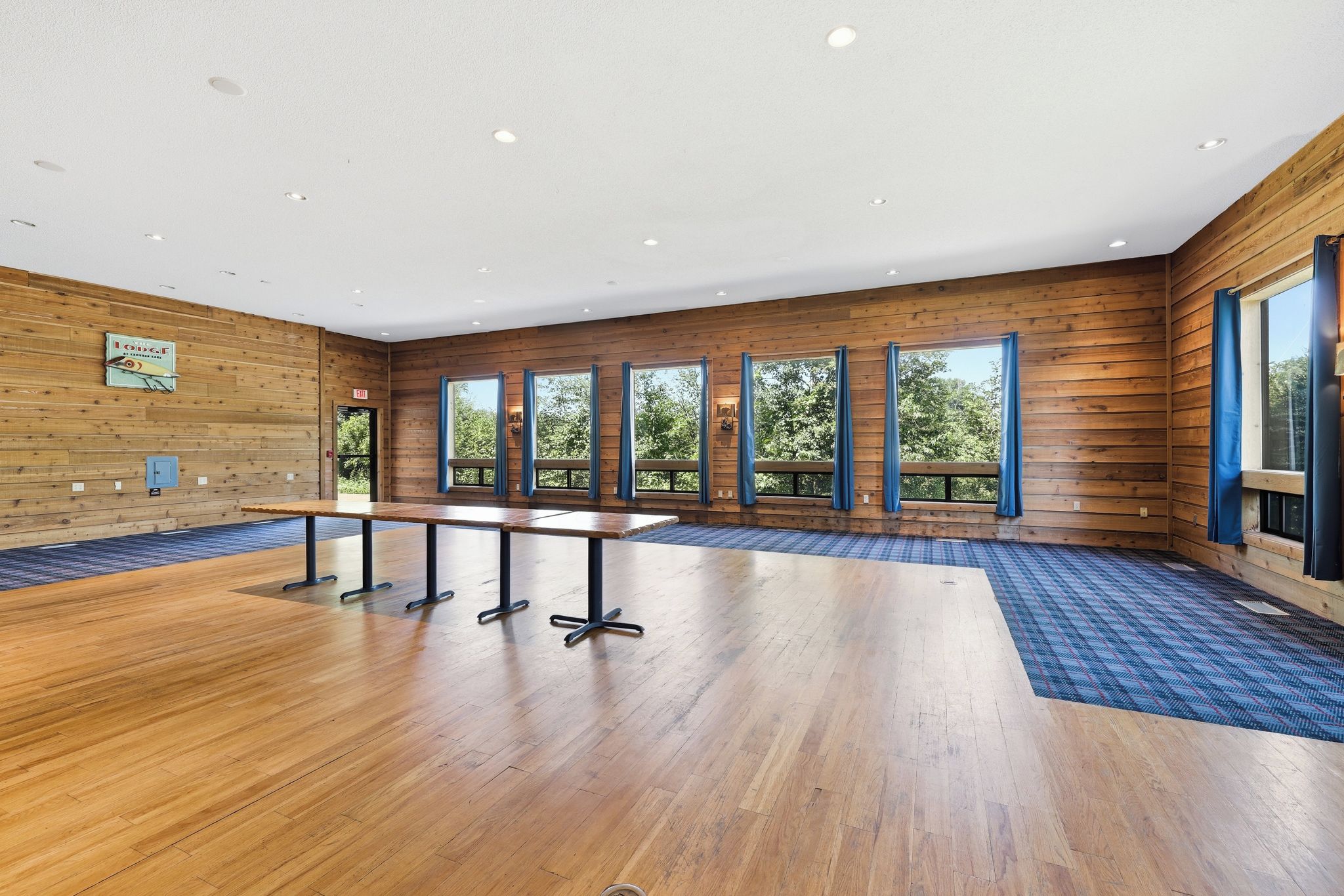 A spacious room with wooden walls, a long table with chairs, large windows with curtains, and a blue patterned carpet.
