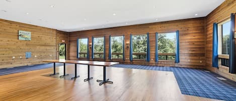 A spacious room with wooden walls, a long table with chairs, large windows with curtains, and a blue patterned carpet.