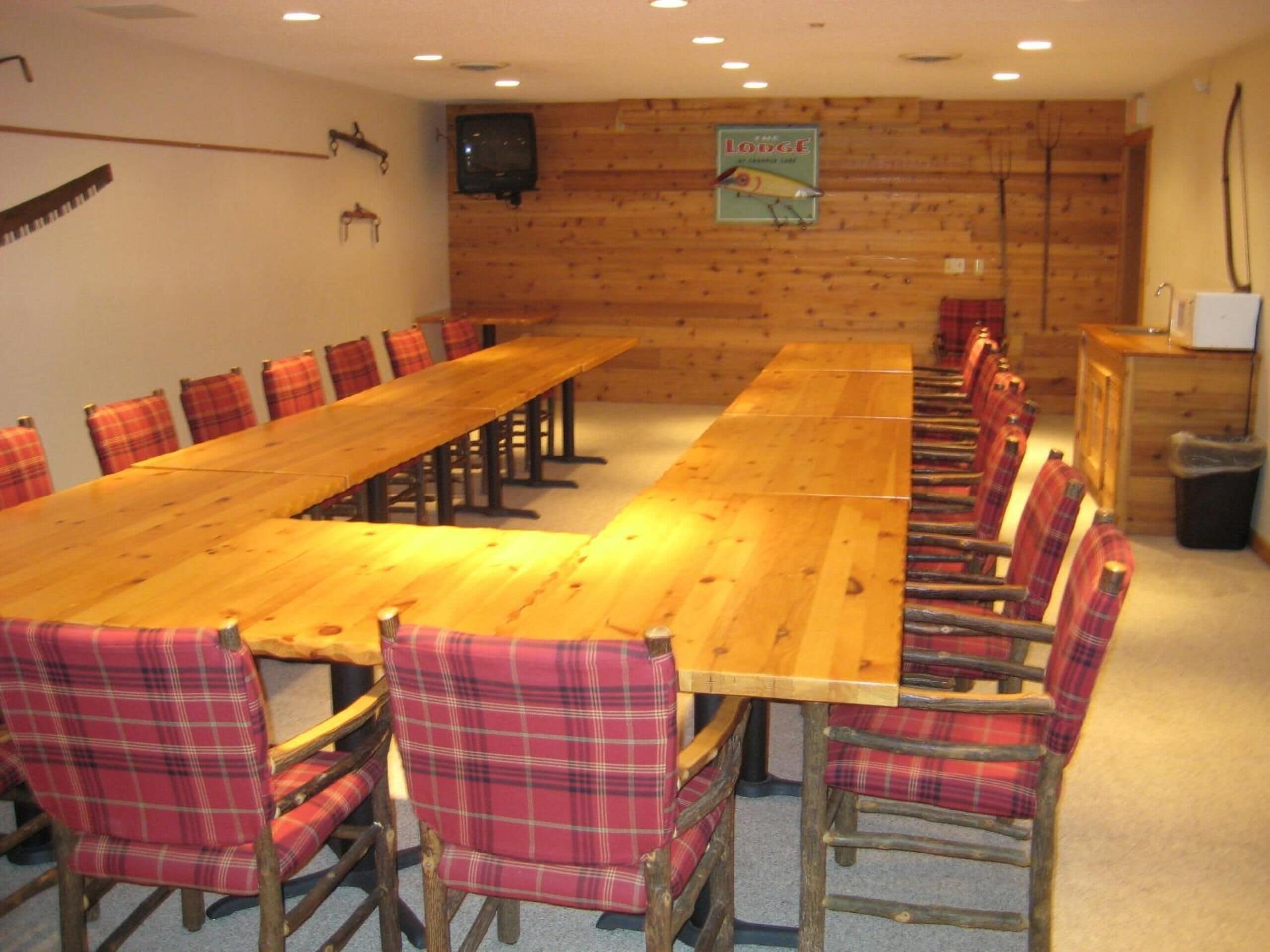 A long wooden table with plaid chairs, a television on the wall, and a sign that reads "The Lodge."